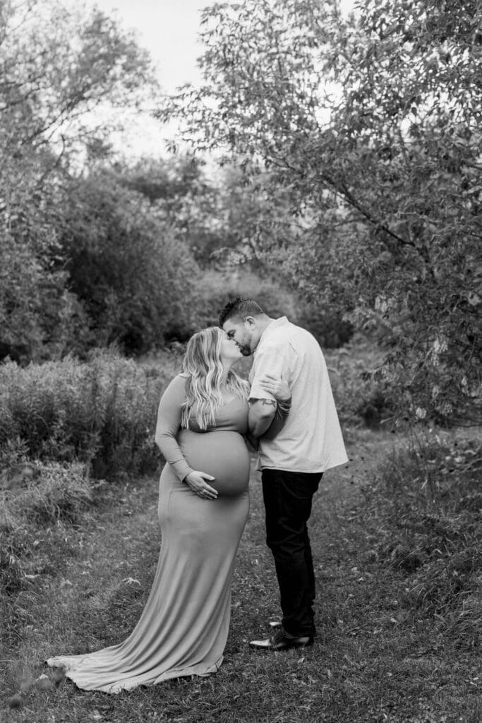 A black-and-white image of a pregnant couple sharing a kiss surrounded by trees and long grass.