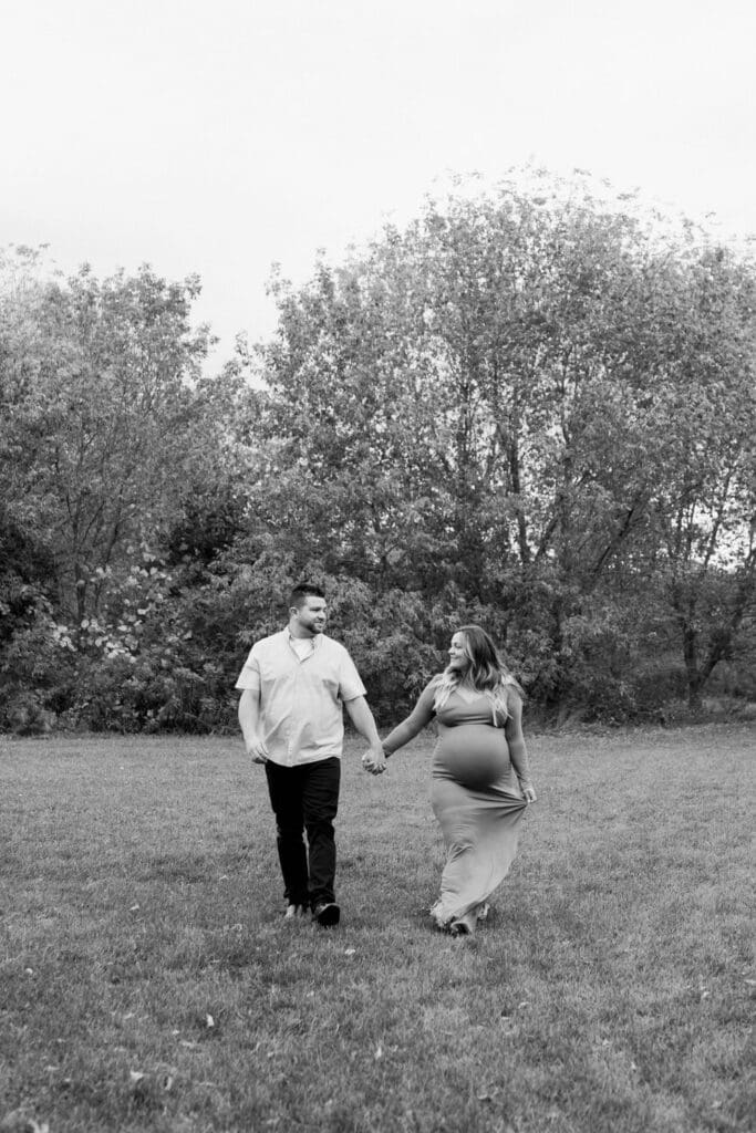 A black-and-white photo of an expecting couple walking hand-in-hand across a grassy field, smiling at each other with trees in the background.