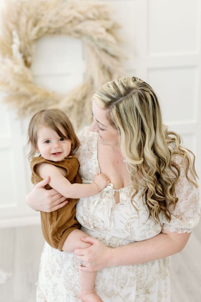 Mother holding smiling toddler in front of a pampas wreath during a family photo session.