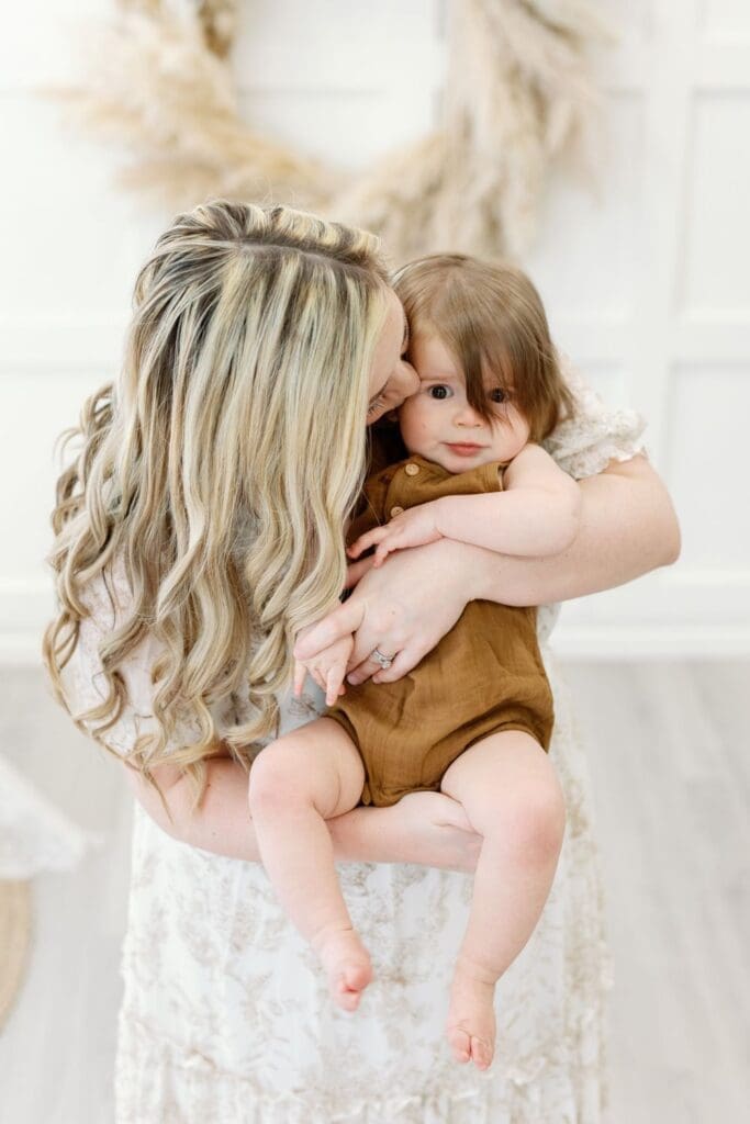 A mother with long blonde curls kisses her baby boy on the cheek while holding him in a neutral-toned studio.