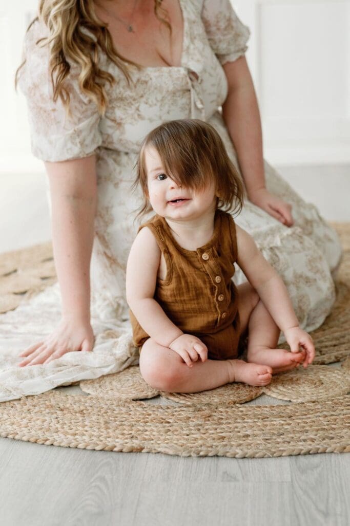 A mother sitting on a woven rug, gazing at her child in a tender moment during a motherhood photography session.