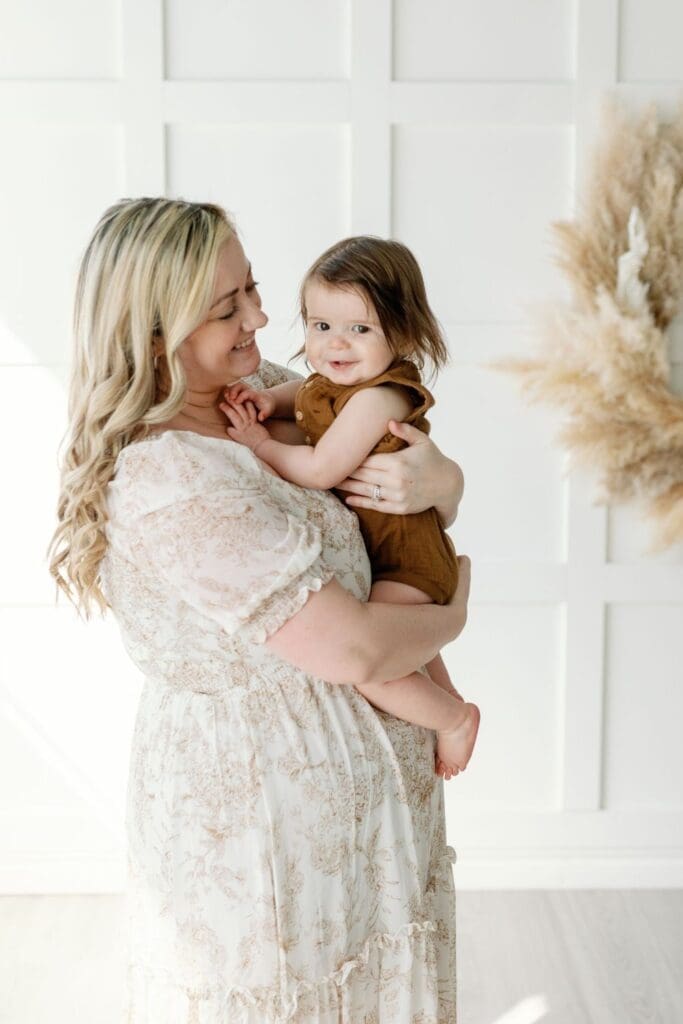 Mother holding smiling toddler in front of a pampas wreath during a family photo session.