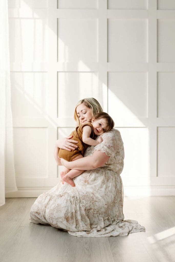 Mother cradling her toddler close in a bright studio with sun spilling through the windows.