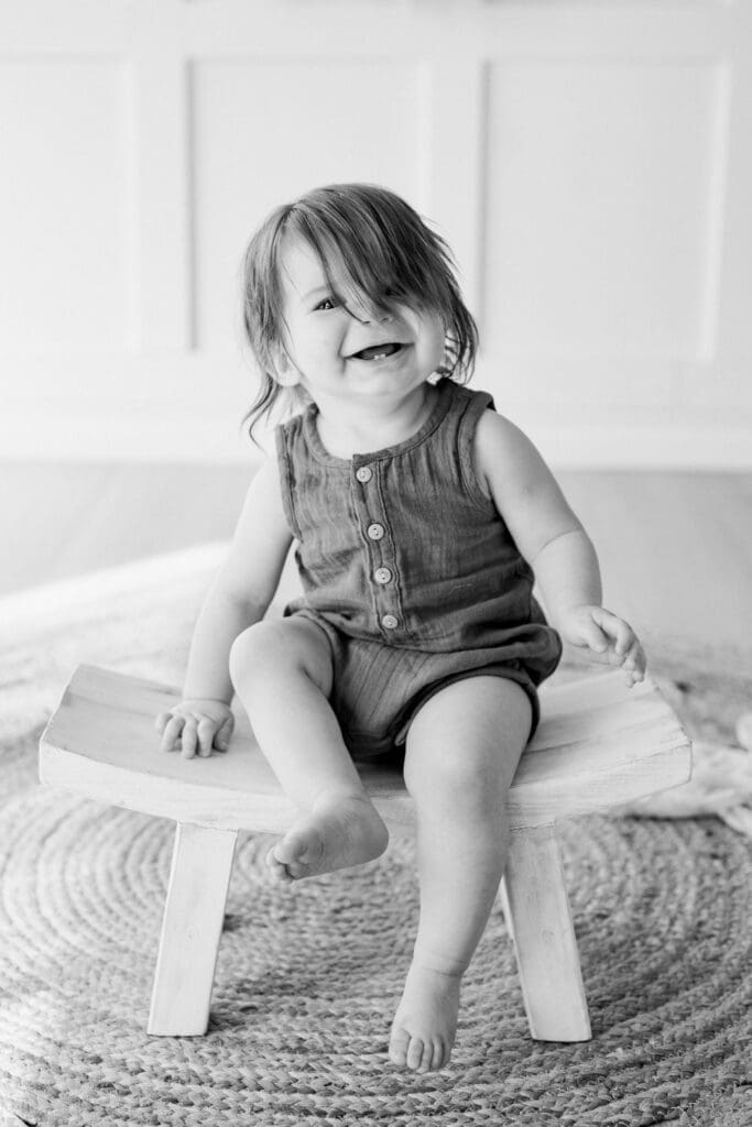 Black and white image of a smiling toddler sitting on a wooden bench during a studio session.