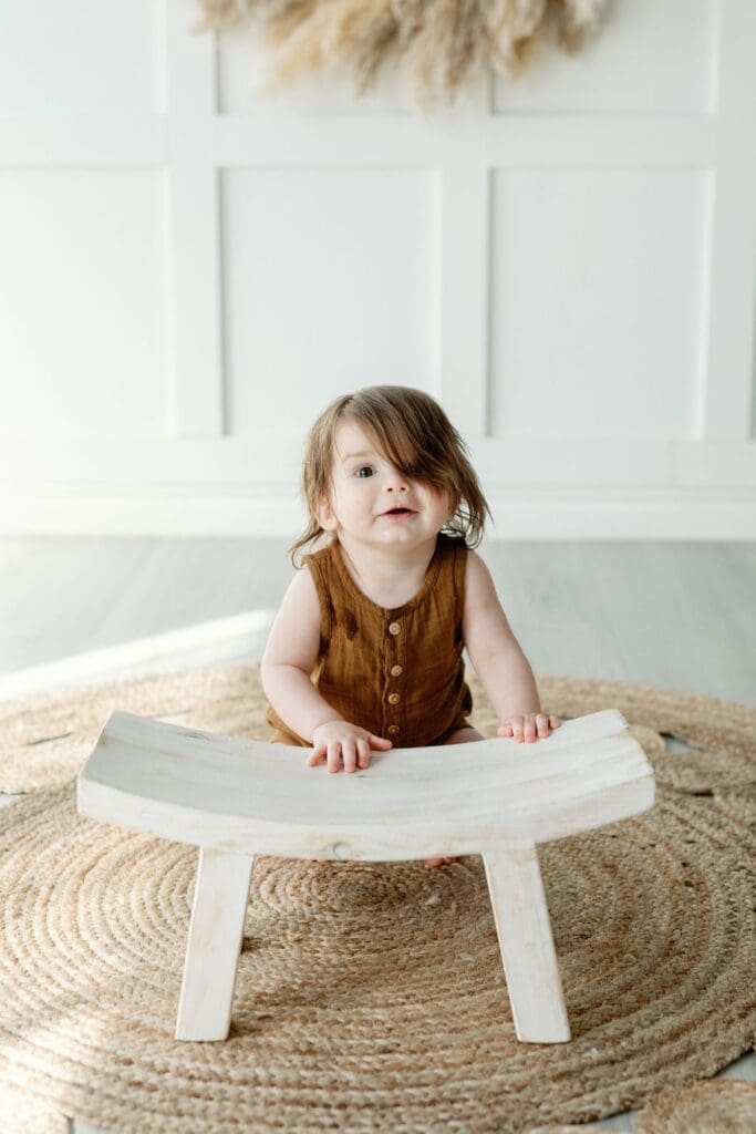 Toddler standing behind a small wooden bench on a jute rug in a white-paneled studio.