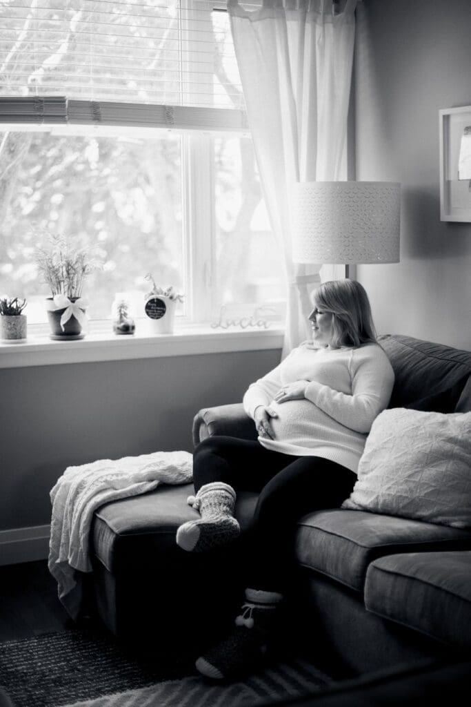 Black and white maternity photo of woman sitting on couch, gazing out snowy window