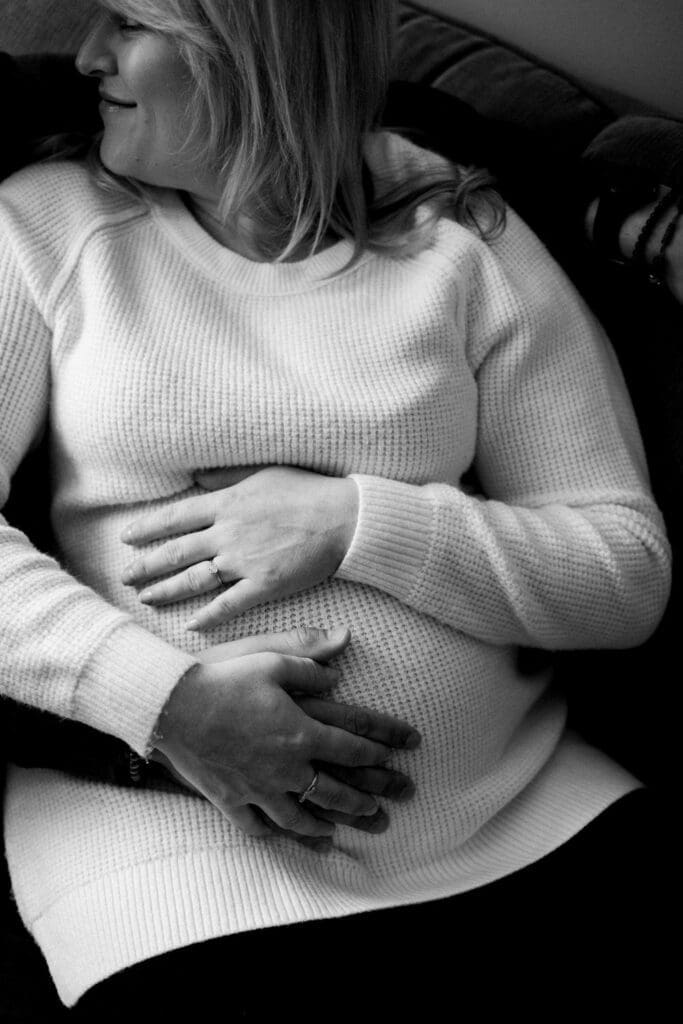 Close-up black and white photo of an expecting couple’s hands resting on the mother’s pregnant belly, both wearing cozy knitwear while sitting on a couch.