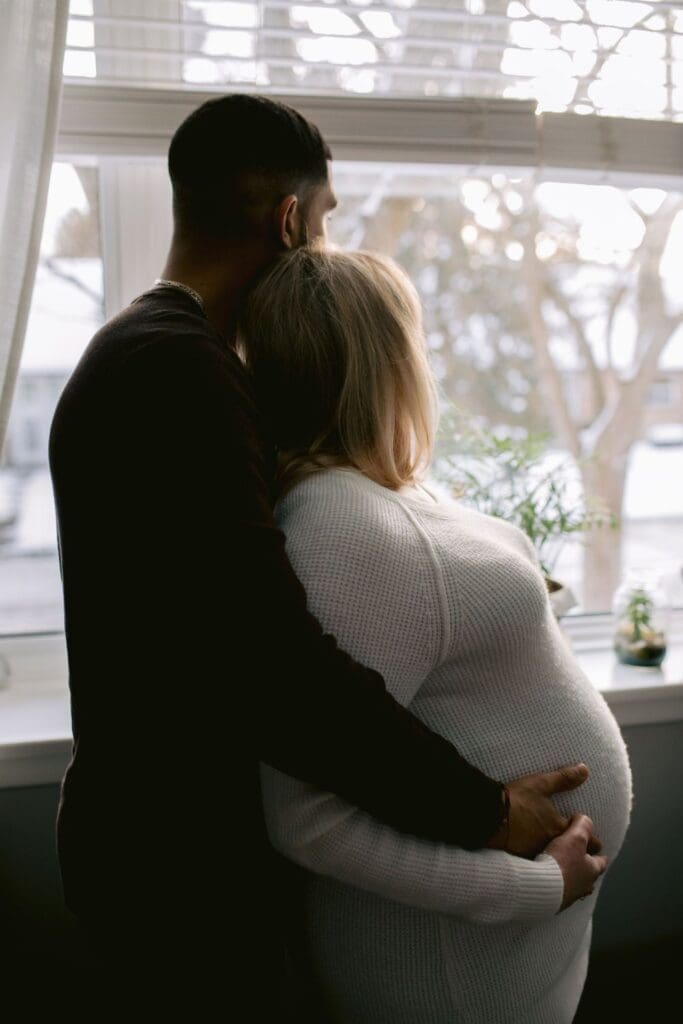 Couple wrapped in each other’s arms looking out the window during indoor maternity session