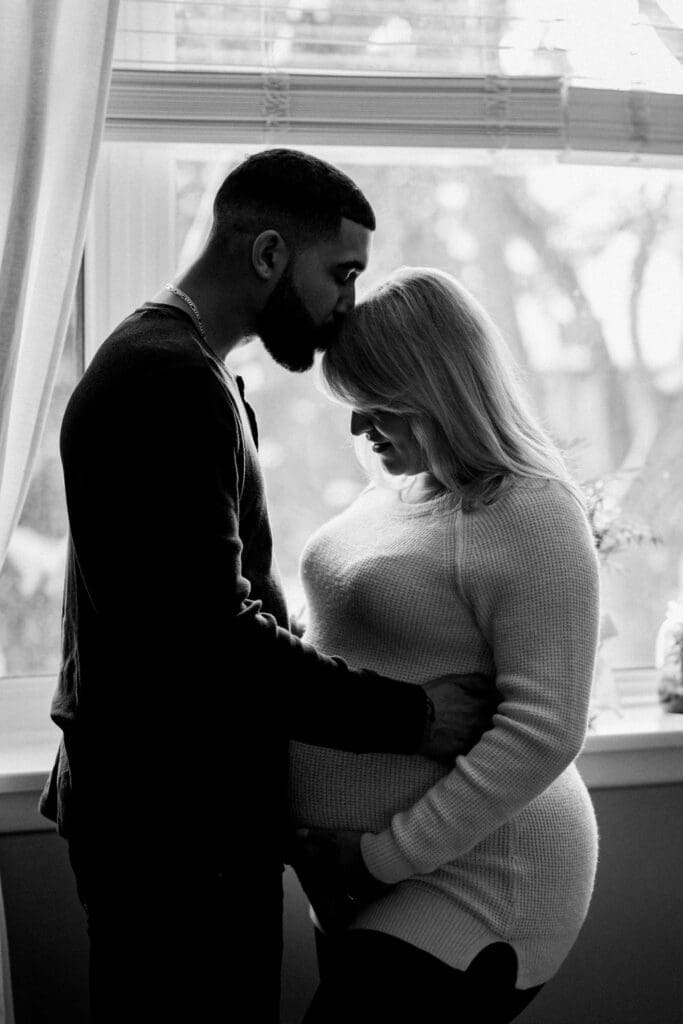 Expectant couple in black and white photo sharing tender moment by the window during maternity session