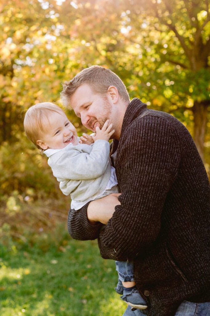Father holding toddler son as they share a joyful moment outdoors with golden fall foliage in the background.