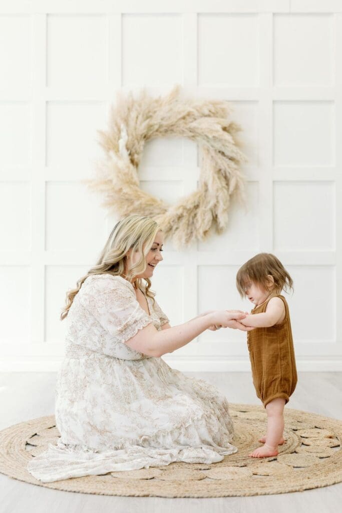 Mother holding her toddler’s hands while sitting on a round jute rug in a white studio.
