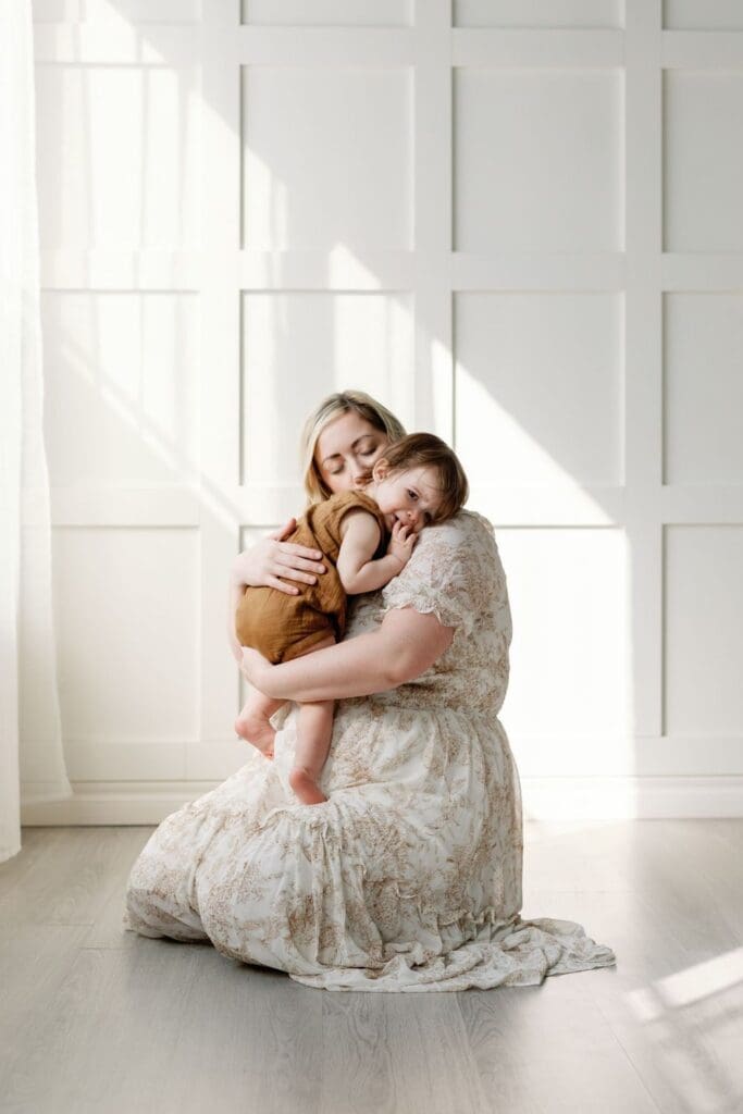 Mother hugging toddler with eyes closed in a white studio during a motherhood photography session.