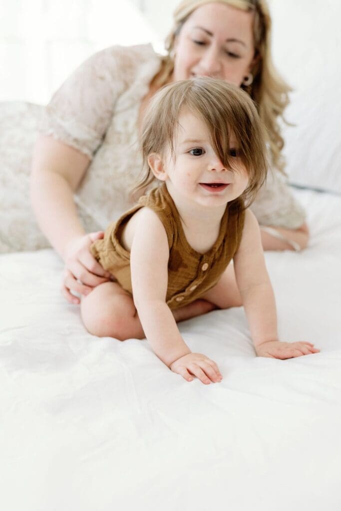 Toddler crawling on a white Sheet while mother smiles in the background during a studio session.